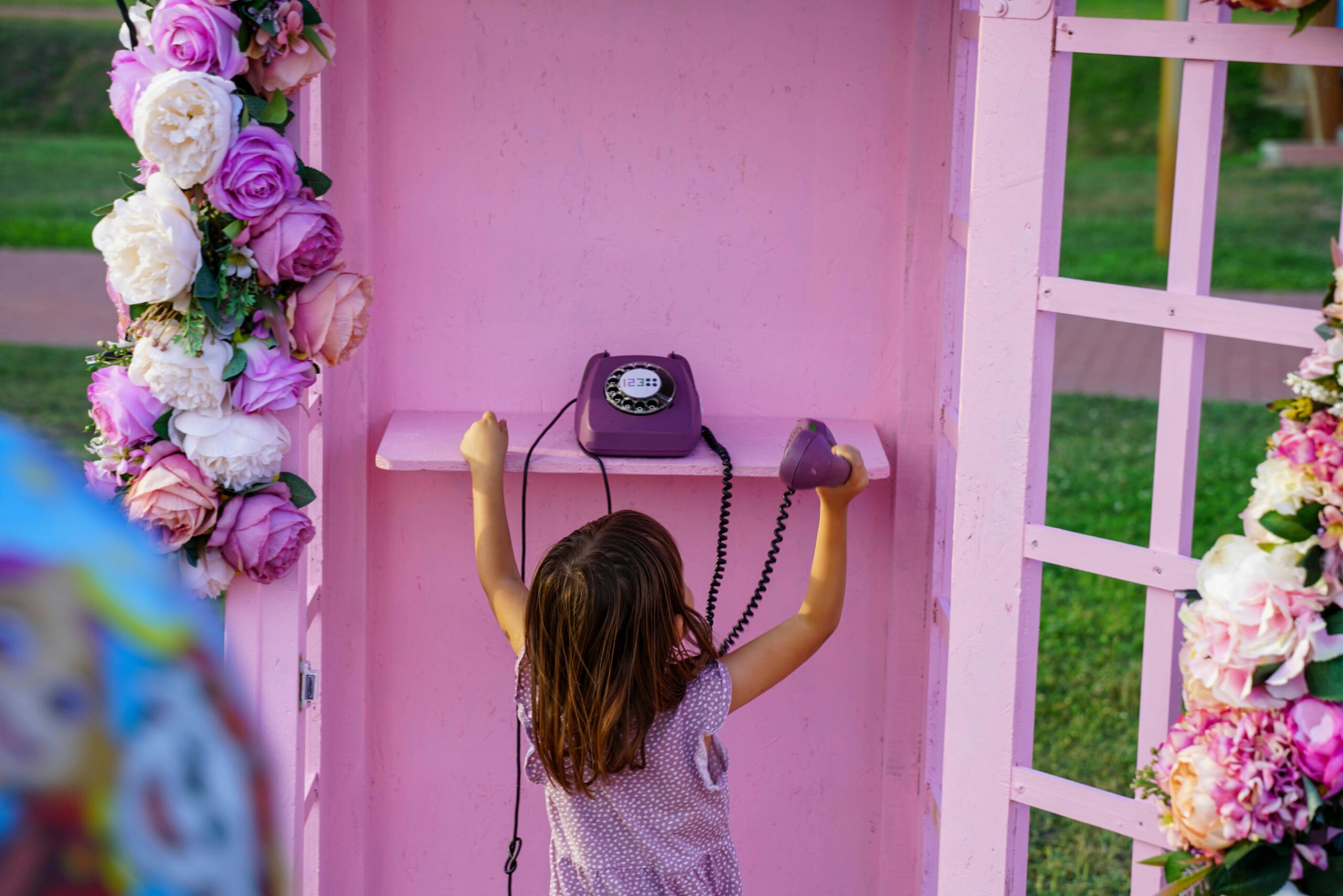 A young girl holds a purple telephone in a pink, flower-decorated booth outdoors.